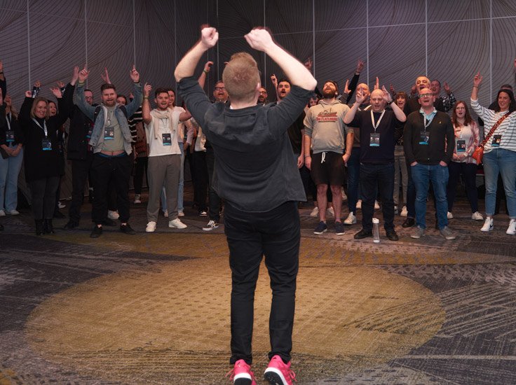 A beatboxer is conducting a beatbox workshop with his hands in the air and a crowd in front copying him